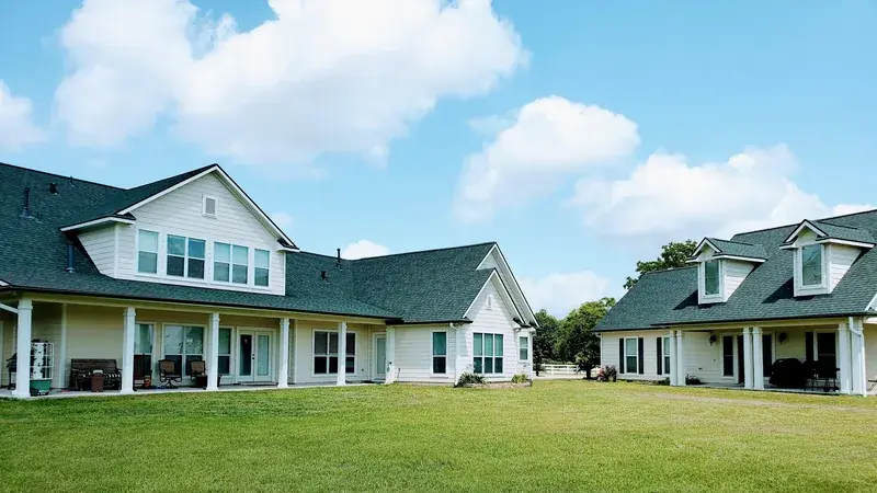 White farmhouse-style home and detached wing with green-gray shingle roofs under a blue sky.