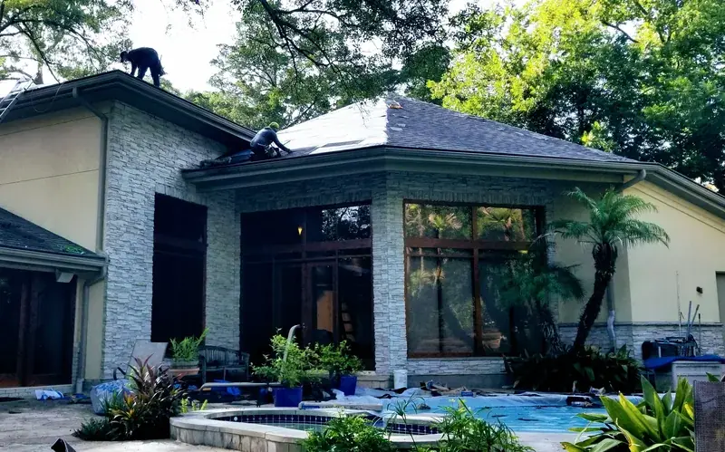 Two roofers laying shingles on a hipped roof above a stone-clad house and pool.