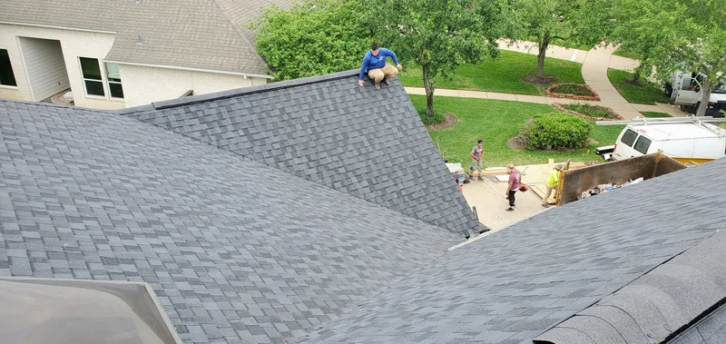 Aerial view of a roofing crew on a grey shingle roof, with a debris dumpster in the driveway below.