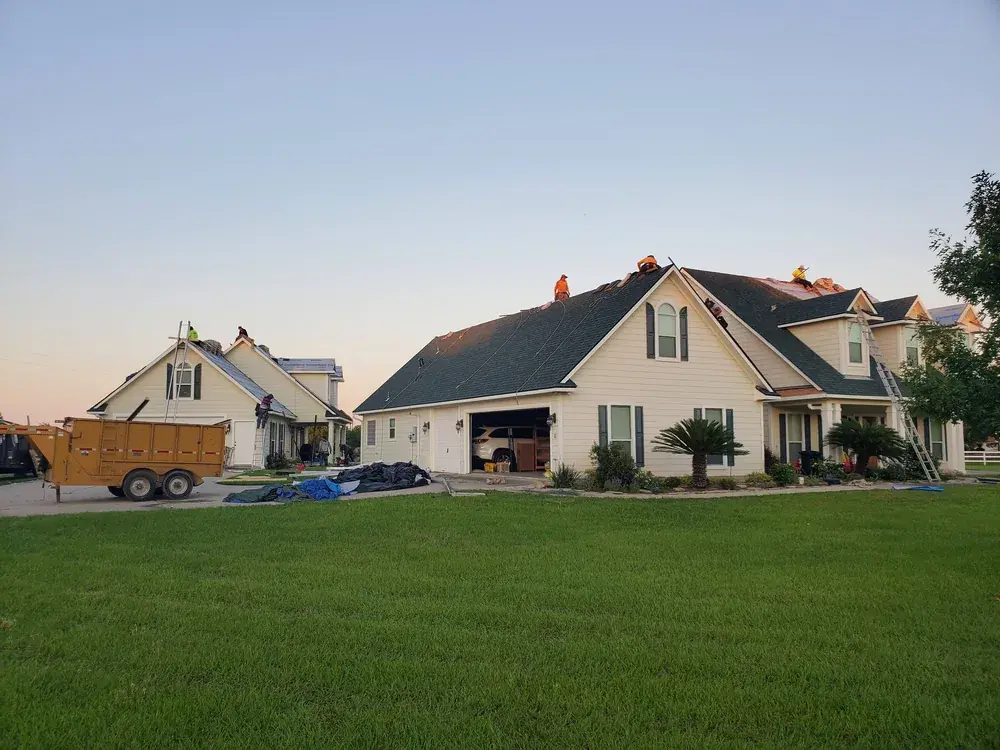 Roofing crew working on the roof of a large single-family home at dusk, with a debris trailer in the driveway.